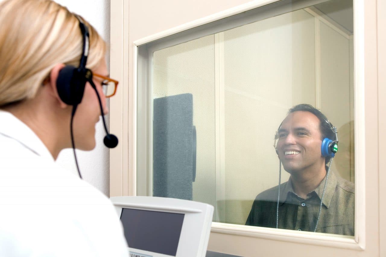 Man receiving a hearing test in a hearing booth looking through a glass window to a female audiologist